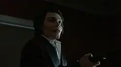 A pale man smiles in a sinister fashion while threatening someone with a gun.