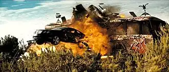 A black car flies through the air out of the side of a graffiti-covered bus that is exploding.