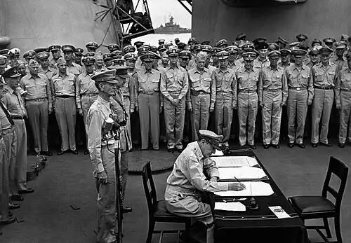 MacArthur is seated a small desk, writing. Two men in uniform stand behind him. A large crowd of men in uniform look on.