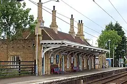 The platform side of the station building as seen from the other platform. The building is a yellow-brick building with red roof tiles and a canopy hanging over the platform. One of the railway tracks and the overhead wires are also visible.