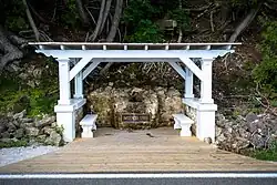 A white, flat-roofed gazebo over a spring, with a plaque located on a rock next to the spring. A short, wide wooden walkway leads from the road to the gazebo.
