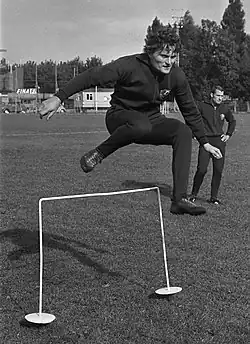 A black-and-white photo of a young man jumping over hurdle.