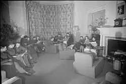 Black and white photograph of men and women sitting on lounge chairs reading newspapers in a large sitting room. Most of the men are wearing military uniform.
