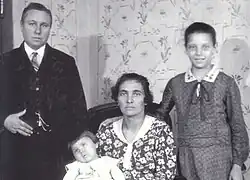A standing woman with very short hair and a suit next to a sitting woman with a small girl and a standing girl