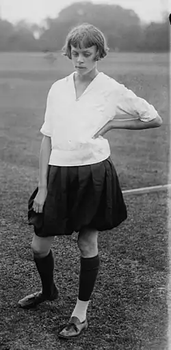 A young white woman with hair cut in a bob with bangs, standing in athletic attire, photographed on a sports field