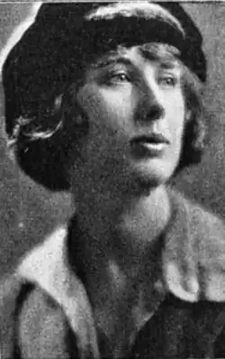 A white woman with hair cut in a bob, wearing a hat and a blouse with an open collar
