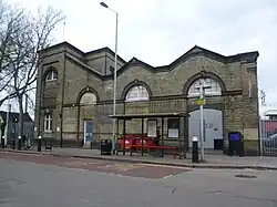 A large yellow-brick building with a bus stop in front as photographed from across the street. Most of the original windows on the facade are bricked in and the doorway is also closed up. A sign appended to the side has a double arrow logo and illegible writing, and overhead line equipment is visible behind the building on the right hand side.