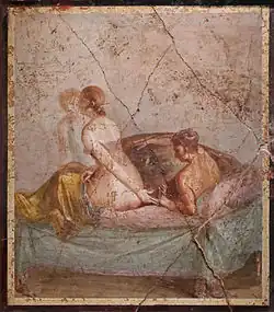 A nude woman, getting up towards the foot of a bed, reaches back towards her male lover, lying down in it.