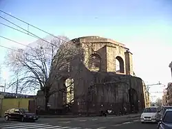 Exterior of a ten-sided ruin called today the Temple of Minerva Medica at the intersection of city streets in Rome showing large arched windows in the drum between engaged buttresses and below polygonal step-rings buttresses for the collapsed dome