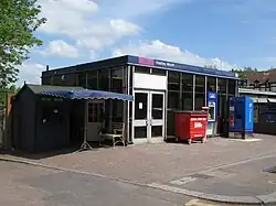 A small cuboid brutalist plastic and glass building as viewed from across the street. In front of the building are a bin, ATM, ticket machine, and flower stall. Above the windows a sign reads "Hadley Wood".