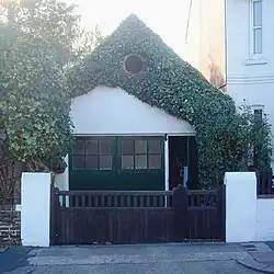 An extremely small workshop adjoining a house and standing behind a wooden gate with white gateposts. It has green double doors with an open door to their right. Above this is a white-painted wall which is heavily draped with ivy, although a circular louvre is clearly visible through this. The roof is steep and pointed.