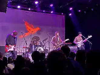 Four rock musicians perform on a stage, illuminated by purple lights and a red cardinal projected on the backdrop