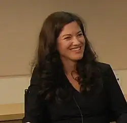 A headshot of a smiling woman in a black jacket with long brown hair.