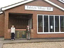 Frontage of a brick-built hall with two young Fulstowians standing by the entrance and the village's war memorial.