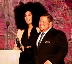 Cher stands on stage with Chaz Bono, smiling. She wears a black outfit with an afro hairstyle, while Chaz wears a suit and tie.