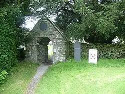 The gate to the churchyard. It is made of cobbled stone and surrounded by overgrown foliage and a low stone wall.