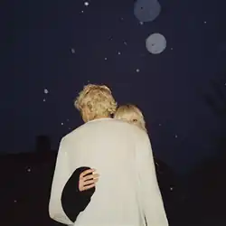 A young man with blonde hair stands with his back facing the camera, while a young woman with blonde hair looks directly into the camera. The woman has her hand around the young man's back.