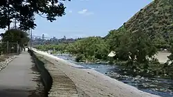 Riverwalk with low wall and natural stream, skyscrapers in far distance