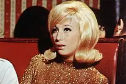 Cher with a voluminous blonde bouffant hairstyle and winged eyeliner, wearing a gold sequined top while seated in a red booth.