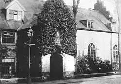 A black and white photograph of the outside of a medieval school hall. The outside is covered in ivy.