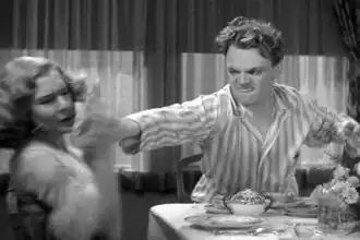Cagney, in striped pajamas, looks angry as he reaches across a breakfast table with the grapefruit in his hand.