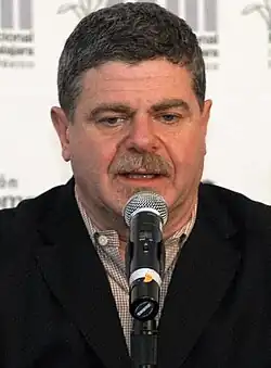 A 56-year-old man with dark gray hair talking into a microphone, looking at something beneath the camera