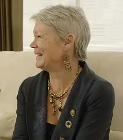 A smiling woman with short grey hair sitting on a beige couch.