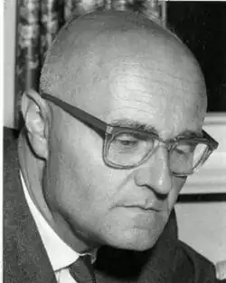 Black and white, three-quarter-profile publicity headshot of a clean-shaven, bespectacled and bald man with serious expression, looking down.