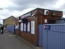 A small cuboid wood panel building wihth a sign above the door that reads "Harringay". It has metal fences on either side with an entrance through a gate. There are signs on the building and fence pointing people to the trains.