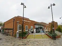 A large single-storey red-brick building slightly above ground level with stairs leading up to it. In the centre is a large glass entrance, and on each side thee are windows with metal letters above that read "Hatfield station". The photo is taken from the pavement in front with lampposts obstructing the building slightly.