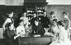 A group of white women posed around a desk and in front of a chalkboard