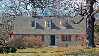 A light yellow one-and-a-half-story wooden house seen from its front, with a large bare tree obstructing part of the view at upper right. It has reddish-orange shutters on the windows and three dormer windows in its shingled roof.