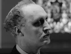 Pale man with dark shadows around his eyes, reaching forward