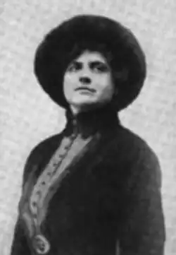 A white woman wearing a dark hat and a matching fitted jacket with a high collar.