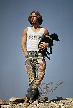 A hitchhiker wearing jeans standing barefoot on rocks while holding a black-colored puppy near U.S. Route 66