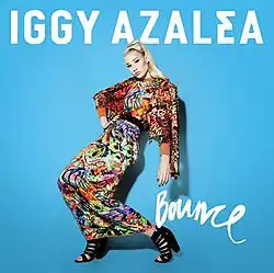 A portrait coloured in light blue, featuring a young blonde woman at its forefront. She is posing in a crouched standing position with her one hand on her hip. She is wearing a traditional Indian sari composed of various colours, and her hair is styled in a high ponytail. Above her in bold white capital font is the name 'Iggy Azalea'. Below her loose arm, in thin cursive white font is the title 'Bounce'.
