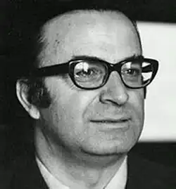 Black and white, three-quarter profile Press shot of a clean-shaven, half-smiling, bespectacled man with a high forehead, short black hair, and sideboards/sideburns