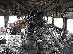 A rail passenger car interior with no upholstery, showing many burn marks and other fire damage. Several long pieces of metal are piled in the center near the back end of the car.