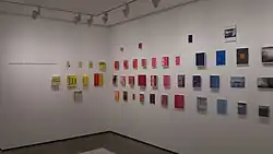 A photograph showing two white walls with a few dozen books nestled inside small transparent display cases mounted to the walls. Books are arranged by color, in a rainbow color order, with yellow on the left and blue on the right end.