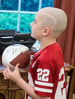 Portrait of Jack Hoffman looking up in the Oval Office. He was then a young, white boy wearing a football jersey and holding a ball signed by President Barack Obama (outside the frame). He has no hair, revealing a scar running across the side of his head to his ear.