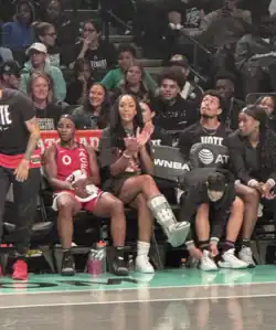 A'ja Wilson wearing street clothes (including a black Dawn Staley T-shirt) sitting on the bench courtside for the Las Vegas Aces. Wilson was out with an injured ankle against the New York Liberty, September 8, 2024, and she is wearing a boot