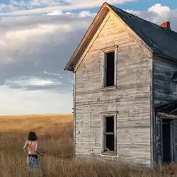 Jane Remover stands alone in a field with their back towards the camera as they face the remnants of a dilapidated house.