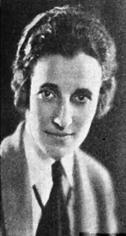 A woman with light skin and wavy dark hair cut short, wearing a menswear-style jacket, shirt, and tie