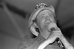 A black and white photograph of a smiling man wearing a cowboy hat and holding a microphone