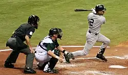 A baseball player in a gray uniform follows through with his swing, while a catcher on the opposing team in a white uniform squats behind him and an umpire in a black uniform stands behind the catcher.