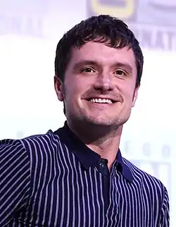 Josh Hutcherson smiling, wearing an open patterned brown and tan flannel, with a brown shirt underneath