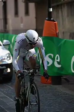 A road racing cyclist wearing a mostly white skinsuit with black, green, and gold trim crouches low on his bicycle while riding out of the saddle. A car following behind him and a roadside barricade are also visible.