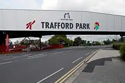 A photograph of a bridge. On the side of the image is, right left to right: a red K (the Kellogg logo), Trafford Park (printed in all capital letters) and a green cockerel (the Corn Flakes logo)