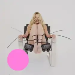 A blonde female sitting in a chair, smiling with her legs drawn up and tied with black ties. She is set against an off-white background, with a large pink dot in the bottom left corner of the image.