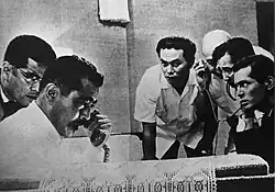 A still from the film depicting Gondo on the phone surrounded by anxious detectives listening to a phone call from the kidnapper